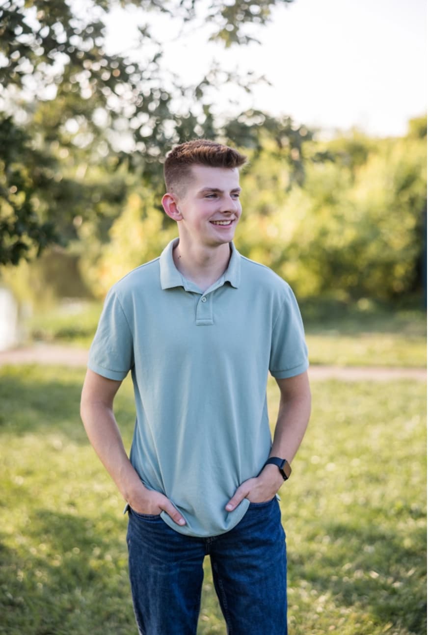 Smiling young man in a light green polo shirt stands outdoors in a sunny park.